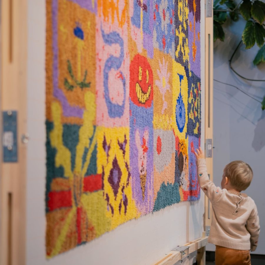 Ett barn sträcker sig mot en stor färgstark tuftad textil.